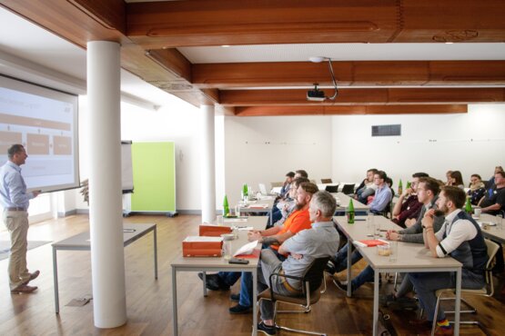 Mann hält vor einem Smartboard einen Vortrag vor einer Gruppe von Menschen