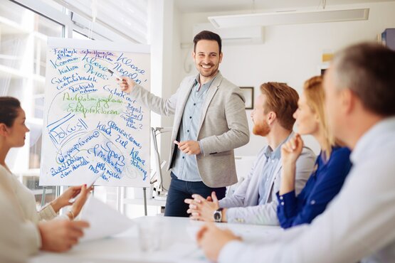Mann deutet vor Publikum mit einem Stift auf ein beschriebenes Whiteboard